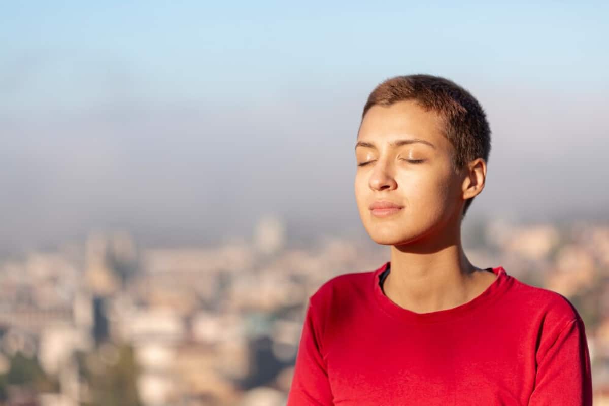 Woman with short hair in a red top, eyes closed, enjoying the sun, with a blurry cityscape in the background.