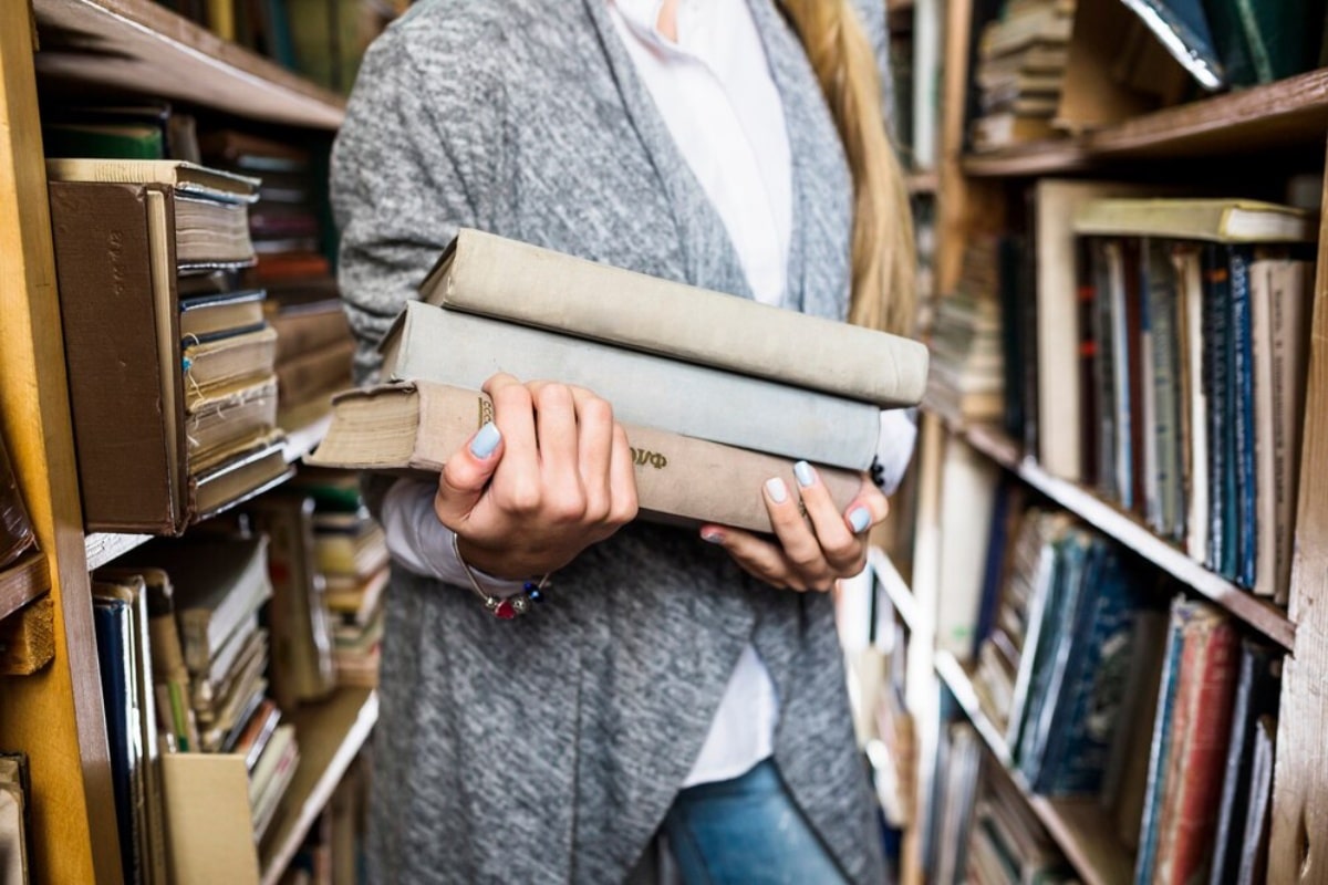 Woman carrying a stack of old books in a narrow, cluttered library aisle.
