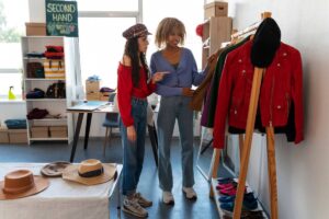 Two women browse clothing items in a cozy second-hand store, surrounded by racks of colorful garments and shoes.