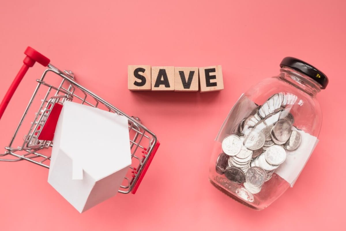 A miniature shopping cart with a house model, wooden blocks spelling save, and a jar of coins on a pink background.