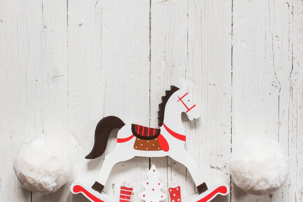A decorated wooden rocking horse among white pom-poms, set against a rustic wooden background, evoking a festive holiday spirit.